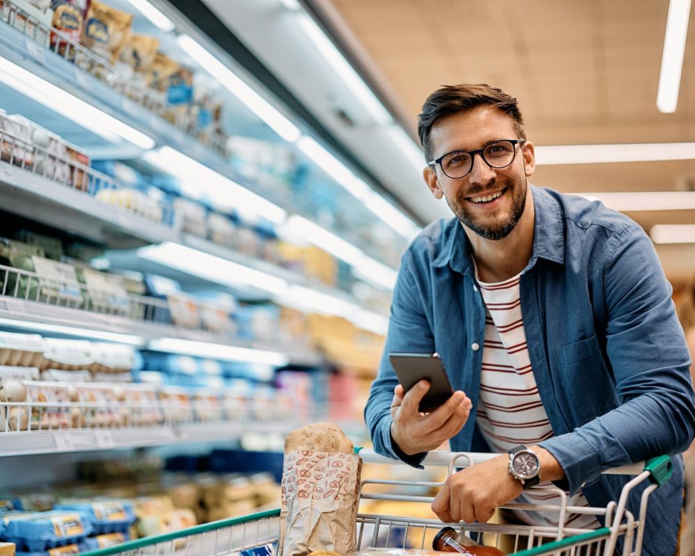 Happy man using mobile phone app while buying groceries in supermarket and looking at camera.